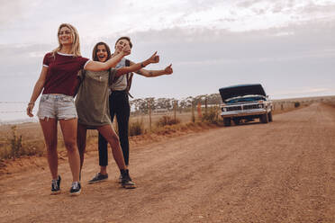 Drei junge Frauen trampen in der Nähe ihres kaputten Autos auf einer Landstraße. Lächelnde Freundinnen gestikulieren für eine Mitfahrgelegenheit auf einer Landstraße mit einem kaputten Auto im Hintergrund. - JLPSF24284