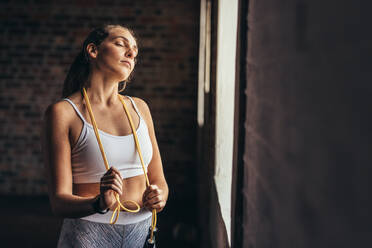 Woman athlete with skipping ropes around her neck standing by gym window. Fitness woman taking break after workout at gym. - JLPSF24317