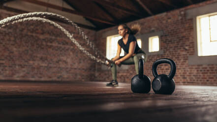 Kettle Bells auf dem Boden vor und eine Frau, die mit dem Battle Rope trainiert. Eine Frau trainiert im Cross-Training-Fitnessstudio. - JLPSF24339