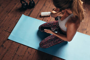 Top view of young woman sitting on yoga mat wearing earphones to listen music on mobile phone after workout. Fitness woman at gym listening to music during break. - JLPSF24343