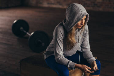 Eine Frau mit Kapuzenpulli sitzt auf einer Box im Fitnessstudio und schaut auf ihr Handy. Eine gesunde Frau ruht sich nach ihrem Training im Fitnessstudio aus und hört Musik mit ihrem Smartphone. - JLPSF24350