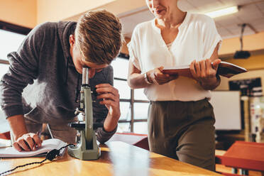 High school student working in a science class with his teacher standing by. Boy looking at slides in biology class through a microscope. - JLPSF24357