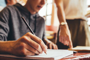 Nahaufnahme eines Teenagers, der während einer Vorlesung am Schreibtisch sitzend Notizen schreibt, während eine Lehrerin auf ein Buch zeigt. Fokus auf die Hand, die im Klassenzimmer in ein Buch schreibt. - JLPSF24363
