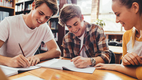 Eine Gruppe von Studenten sitzt am Tisch in der Bibliothek und lernt zusammen. Glückliche junge Freunde arbeiten an einem Highschool-Projekt. - JLPSF24368