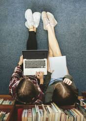 Top view of two female students sitting on floor by a bookshelf and working on a laptop. Two girl school students studying together in library. - JLPSF24369