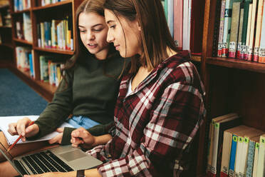 Studentinnen sitzen in der Bibliothek mit einem Buch und einem Laptop und lernen zusammen. Zwei Studenten arbeiten an einer Schulaufgabe in der Bibliothek. - JLPSF24371