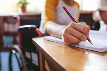 Nahaufnahme einer Studentin, die im Rollstuhl sitzend am Schreibtisch Notizen macht. Fokus auf die Hand, die mit einem Bleistift in ein Notizbuch schreibt. - JLPSF24376
