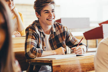 Teenage boy smiling during lecture in high school classroom. Male student making notes and smiling during a period in college classroom. - JLPSF24393