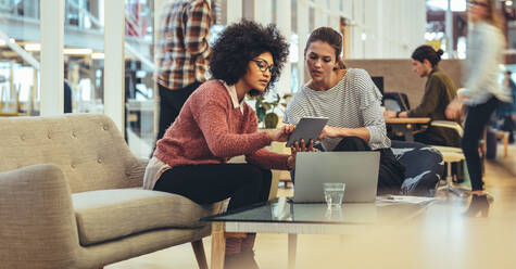 Unternehmerinnen sitzen in einer Lounge am Arbeitsplatz und tauschen mit Tablet und Laptop Ideen aus. Geschäftsfrauen in einer Besprechung, die die Arbeit im Büro besprechen. - JLPSF24405