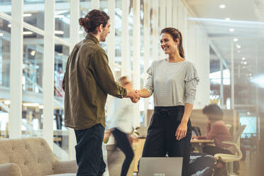 Eine Unternehmerin begrüßt einen Geschäftsmann, der im Büro steht. Ein Geschäftsmann schüttelt die Hände mit einem Mitarbeiter am Arbeitsplatz. - JLPSF24408