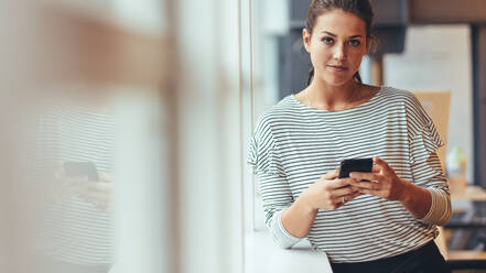 Geschäftsfrau, die am Fenster steht und ihr Handy überprüft. Frau, die eine Arbeitspause einlegt, steht mit Handy in der Hand. - JLPSF24412