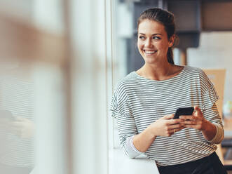 Fröhliche Unternehmerin, die an einem Fenster steht und hinausschaut. Geschäftsfrau, die mit einem Mobiltelefon in der Hand im Büro steht. - JLPSF24414