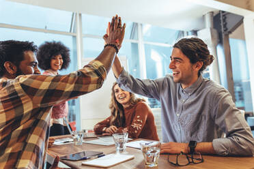 Coworkers sitting in conference room celebrating success. Businessman giving a high five to his coworker while their female colleagues look on. - JLPSF24423