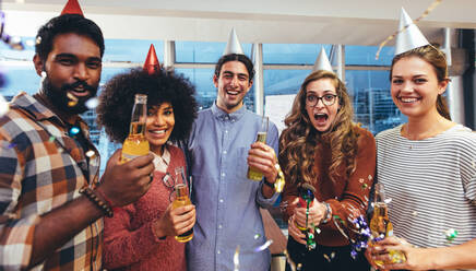 Smiling men and women in party caps and holding beer enjoying a party. Coworkers having fun at a party in office to celebrate success. - JLPSF24427