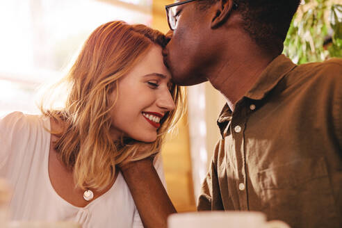 Afrikanischer Mann küsst seine Freundin auf die Stirn. Verliebtes gemischtrassiges Paar bei einem Date im Coffee Shop. - JLPSF24434
