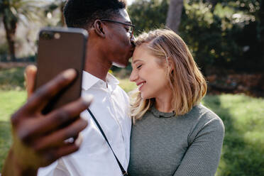 Verliebtes Paar, das ein Selfie mit einem Smartphone im Freien macht. Ein Mann küsst seine Freundin auf die Stirn, während er ein Selbstporträt macht. - JLPSF24448