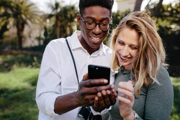 Lächelndes gemischtrassiges Paar, das zusammen steht und sich die Bilder auf dem Smartphone ansieht. Junger Mann und Frau, die sich die Fotos auf dem Mobiltelefon ansehen. - JLPSF24450
