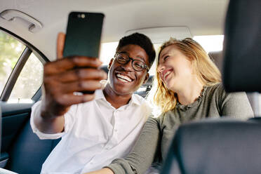 Fröhliches gemischtrassiges Paar sitzt auf dem Rücksitz eines Autos und macht ein Selfie mit dem Handy. Lächelnder Mann mit Freundin, die ein Selfie machen, während sie mit einem Auto unterwegs sind. - JLPSF24453