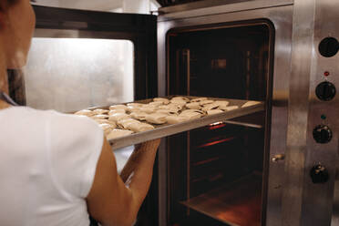 Bäckerin schiebt ein Backblech mit Brötchen in den Ofen. Frau backt Brötchen in einer Bäckerei. - JLPSF24456