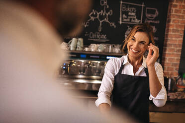 Lächelnde Frau mit Schürze bei der Arbeit in einer Bäckerei. Bäckerin bei der Arbeit mit Blick auf einen Mann vor ihr. - JLPSF24457