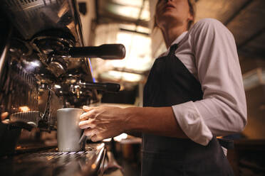 Frau, die in einem Café arbeitet und eine Schürze trägt, macht Kaffee in einer Maschine. Barista macht Kaffee in einer Kaffeemaschine. - JLPSF24461