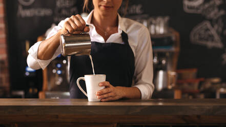 Ausschnitt einer weiblichen Barista, die eine Tasse Kaffee zubereitet, während sie hinter dem Tresen eines Cafés steht. Junge Frau gießt Milch in eine Tasse Kaffee. - JLPSF24463