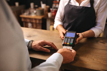 Abgeschnittene Aufnahme eines männlichen Kunden, der in einem Café mit einer Kreditkarte für einen Kaffee bezahlt. Eine Barista-Frau hält ein Kreditkartenlesegerät in der Hand, während ein Mann auf dem Tresen des Cafés die Zahlung vornimmt. - JLPSF24466