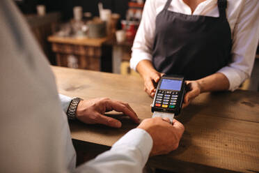 Cropped shot of male customer paying for coffee by credit card at cafe. Woman barista holding a credit card reader machine with man doing payment on cafe counter. - JLPSF24466