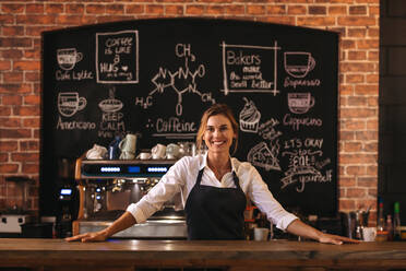 Portrait of confident female barista standing behind counter. Woman cafe owner in apron looking at camera and smiling. - JLPSF24467