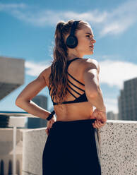 Fit young woman in sportswear and headphones standing outdoors. Female athlete with headphones standing outside and looking away. - JLPSF24472