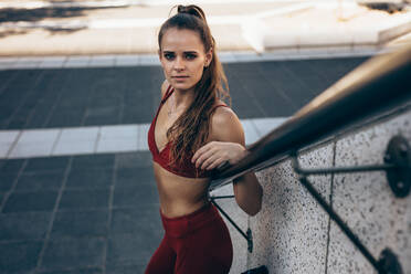 Portrait of a fit woman standing outdoors by a stairway. Fitness female relaxing after workout outdoors. - JLPSF24483