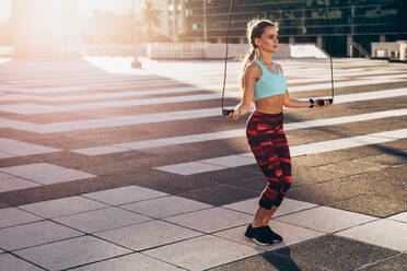 Full length of woman in sportswear skipping ropes outdoors. Female athlete doing cardio workout in morning. - JLPSF24506