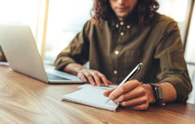 Unternehmer macht Notizen an seinem Schreibtisch im Büro sitzend. Geschäftsmann sitzt mit einem Laptop auf dem Tisch und schreibt Notizen in ein Buch. - JLPSF24513