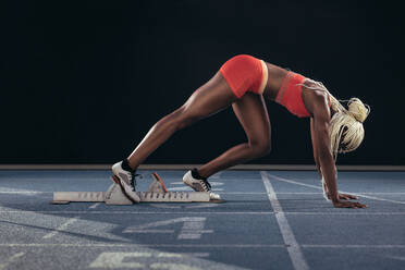 Female athlete taking position on her marks to start off the run. Side view of female runner getting ready to race at the start line on running track on a black background. - JLPSF24548