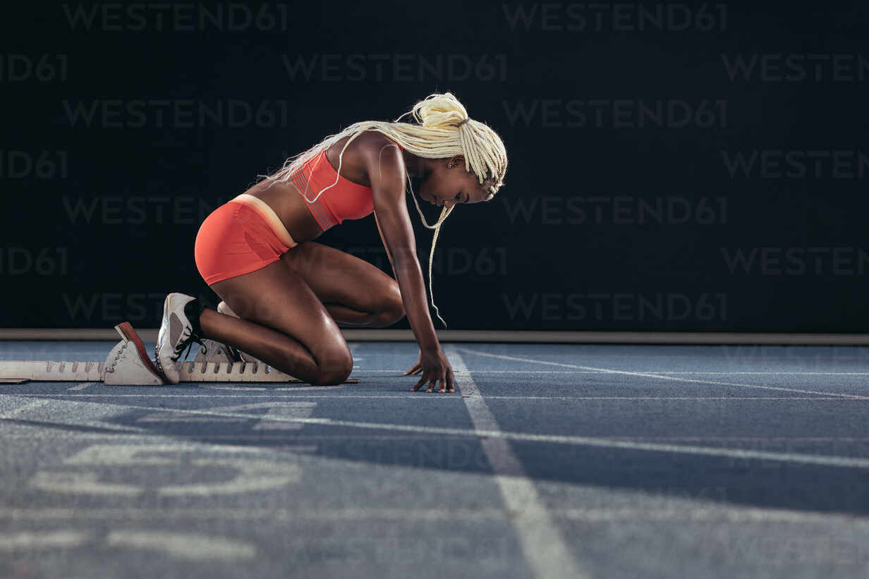 Female athlete taking position on her marks to start off the run. Side view  of female runner sitting at the start line on running track on a black  background. stock photo