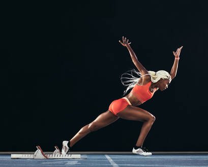 Side view of a female athlete starting her sprint on an all-weather running track on a black background. Runner using starting block to start her run on a running track. - JLPSF24553