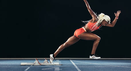 Seitenansicht einer Athletin, die ihren Sprint auf einer Allwetter-Laufbahn vor schwarzem Hintergrund beginnt. Läuferin mit Startblock, um ihren Lauf auf einer Laufbahn zu beginnen. - JLPSF24554