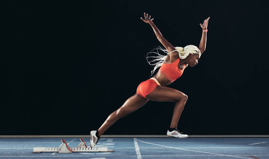 Seitenansicht einer Athletin, die ihren Sprint auf einer Laufbahn vor einem schwarzen Hintergrund beginnt. Läuferin, die ihren Lauf auf einer Laufbahn mit einem Startblock beginnt. - JLPSF24555