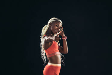 Lächelnde Sportlerin in Fitnesskleidung, die mit der Hand eine Geste macht und mit den Augen zwinkert. Porträt einer Sprinterin in fröhlicher Stimmung beim Training. - JLPSF24564