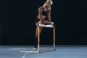 Eine Sportlerin steht auf einer Laufbahn und stützt ein Bein auf eine Hürde. - JLPSF24572