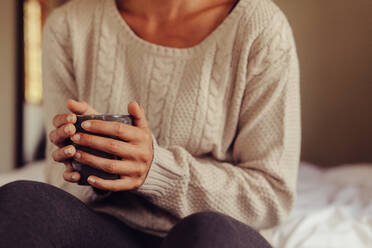 Close up of woman wearing sweater holding a cup of coffee while sitting on bed. Cropped shot of woman having morning coffee in bed. Focus on hands holding a coffee cup. - JLPSF24590