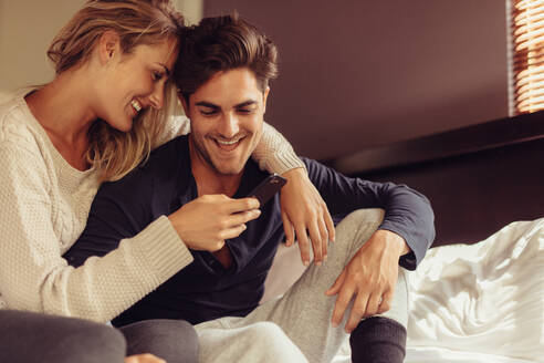 Romantisches Paar sitzt auf dem Bett und schaut auf das Handy. Liebender Mann und Frau lächelnd mit einem Handy. - JLPSF24593