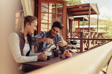 Ein Paar sitzt vor einer Ferienhütte und trinkt Kaffee. Ein Mann gießt Kaffee in eine Tasse, während seine Freundin daneben sitzt. - JLPSF24597