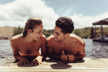 Porträt eines glücklichen jungen Paares auf einem Steg am See, das sich anschaut und lächelt. Junger Mann und Frau auf einem Holzsteg nach einem Bad im See. - JLPSF24612