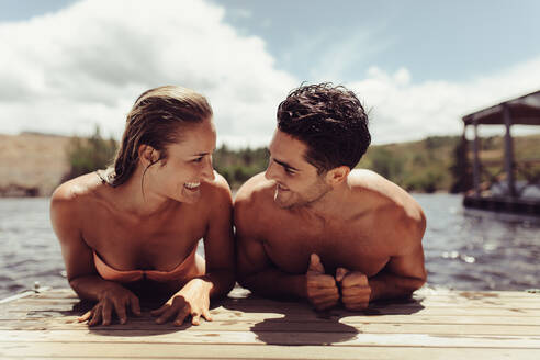 Portrait of happy young couple hanging on pier by the lake looking at each other and smiling. Young man and woman on wooden jetty after a swim in lake. - JLPSF24612