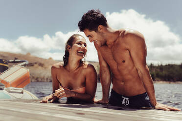 Porträt eines fröhlichen Paares, das sich im Urlaub amüsiert. Junger Mann und Frau klettern nach einem Bad im See auf den Steg. - JLPSF24614