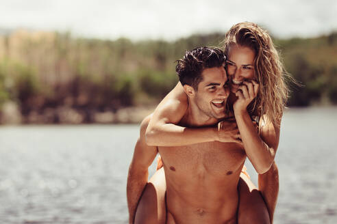 Handsome young man carrying his beautiful girlfriend on his back by the lake. Couple enjoying their holidays outdoors. Piggyback ride fun. - JLPSF24618