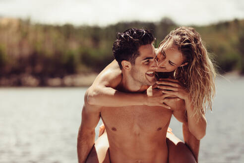 Porträt eines romantischen jungen Mannes, der seine Freundin huckepack nimmt. Junges Paar, das sich am See vergnügt. - JLPSF24619