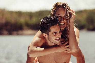 Porträt eines glücklichen jungen Mannes, der seine Freundin huckepack nimmt. Fröhliches junges Paar, das seinen Urlaub am See genießt. - JLPSF24620