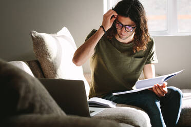 Male student studying on laptop and books sitting at home. Young man preparing for exam studying at home sitting on a couch. - JLPSF24625